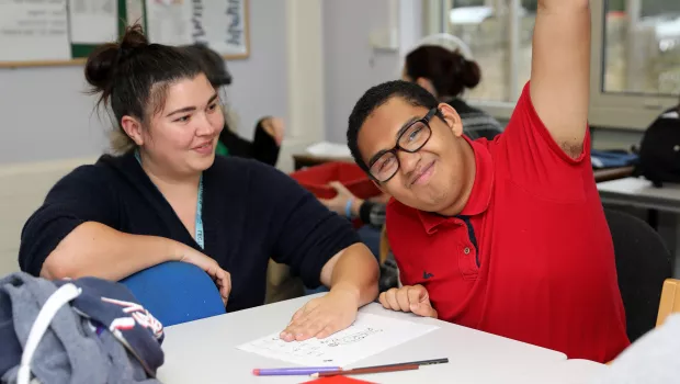 Male student in a red t-shirt, sat at a school desk with a support worker,  looking directly at the camera, smiling with his hand raised.  