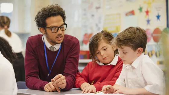 A man in glasses sits with two young students writing in a book in school.