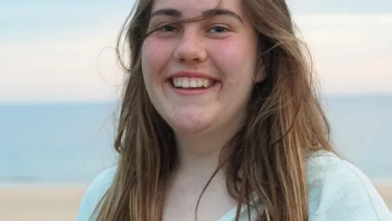 Girl with fair hair smiles at camera, with the sea and sand in the background 