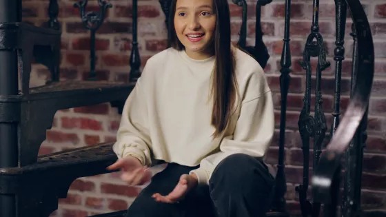 A young woman is sat on a spiral staircase talking to someone off camera