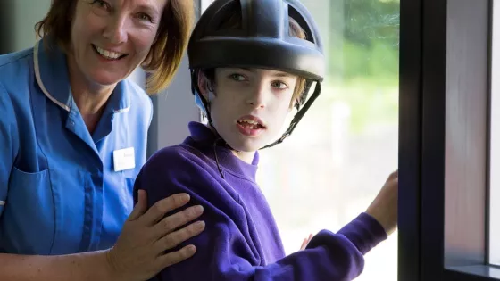 Nurse and young boy who is wearing a protective helmet stand in by the window facing the camera 
