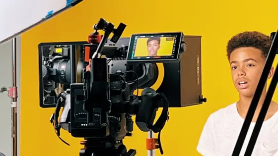 Photograph of teenager on a film set.  The back drop is yellow and he is sat on a stool talking to the camera. 