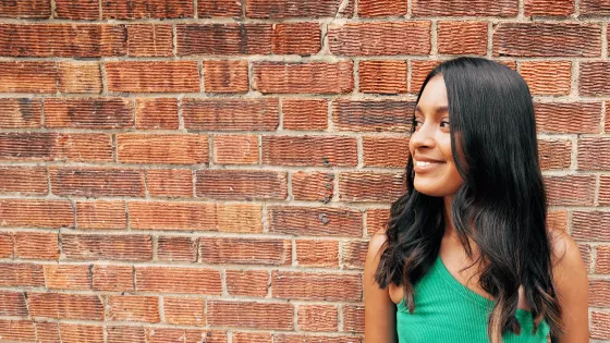 Photograph of young woman stood against a brick wall smiling at someone off camera 