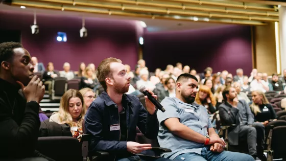 Photograph of three young men the audience of a conference, one holding a mic 