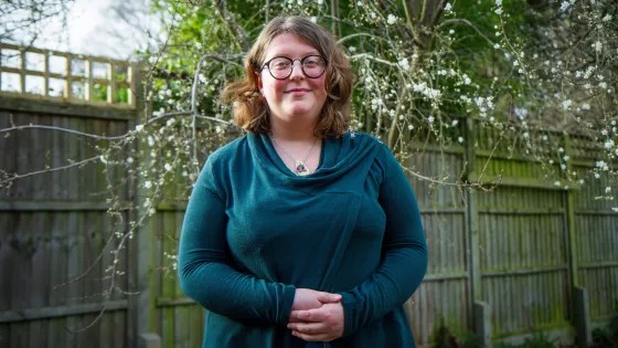 A young woman in a teal jumper and glasses smiles outside for a photo.