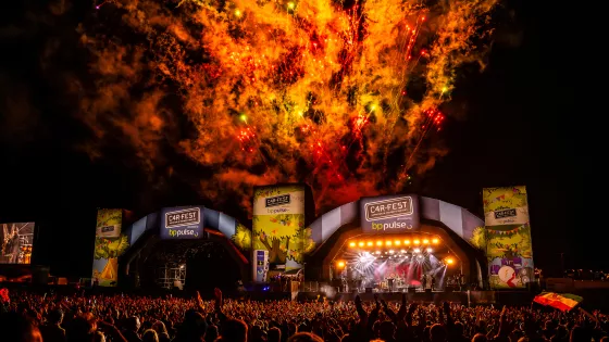 Fireworks light the night sky behind the concert stage and large crowd at Car-Fest. 