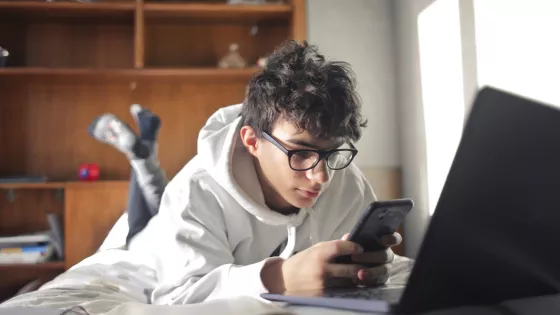 Young boy lying on his bed looking at his phone and laptop