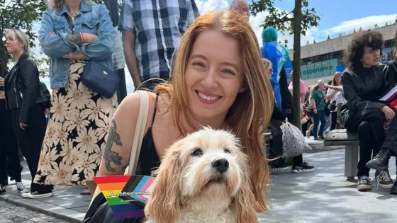 A young woman smiles with a dog while holding a rainbow pride and trans pride flag.