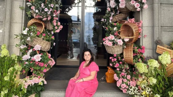 A young woman, Beth, sits surrounded by pink flowers in a doorway.