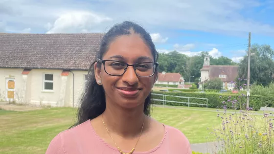 Tee, a young woman in glasses, smiles at camera outside in a rural area.