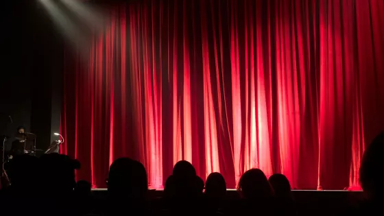The silhouette of an audience in shadow look to a red stage curtain.
