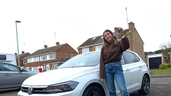 A young woman stands in front of her new car, holding up her keys.