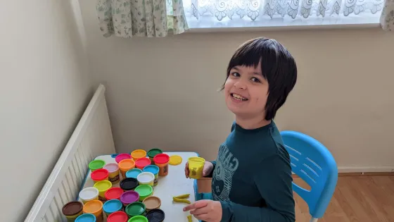 A young boy sits smiling before a table with lots of colours of play dough pots.