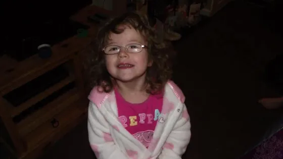 Young girl with curly hair, smiling at the camera, wearing a pink Peppa Pig t-shirt.