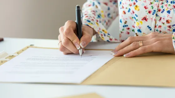 A lady wearing a floral shirt signs a Will document at a desk.