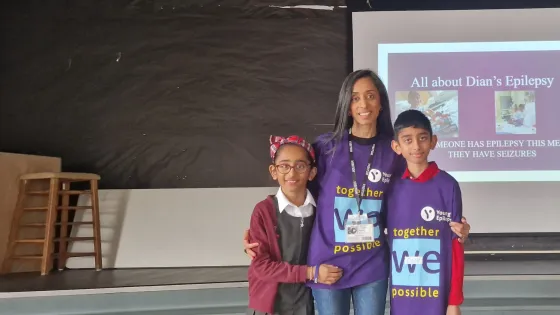 A mother stands with her two children in Young Epilepsy t-shirts in front of a screen.