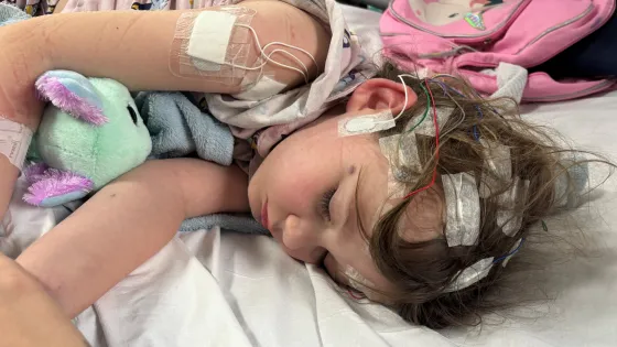 A young girl sleeps in a hospital bed with lots of tubes and patches on her head and arms.