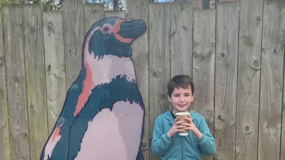 A young boy, Tate, stands beside a fence with a painting of a penguin.