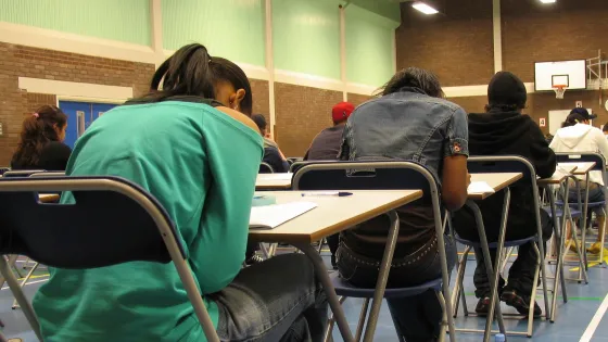 A group of students work on exams, heads down, in a school hall.
