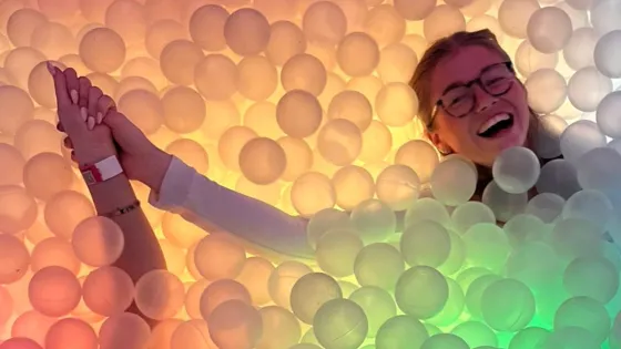 Two women lie down in a rainbow-themed ball pit.