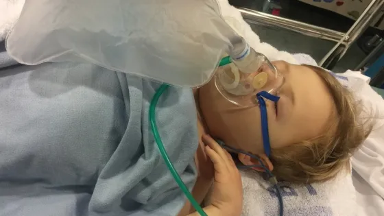 A boy lies in a hospital bed with a breathing mask.