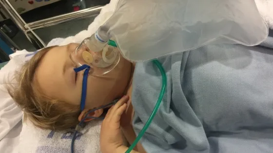 A boy sleeps with an oxygen mask in a hospital bed.