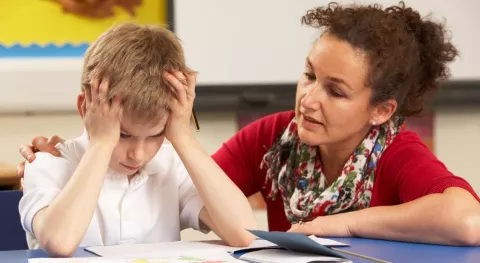 An educator speaks gently to a young student, who is holding his hair in frustration.