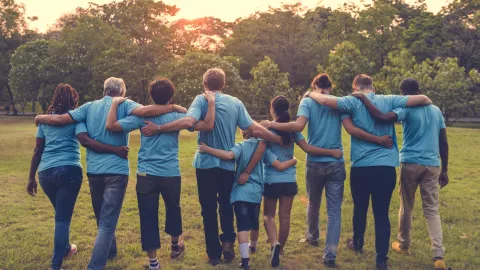 Nine young people with matching blue t-shirts hold shoulders and watch the sun set behind a row of trees.