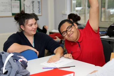 Male student in a red t-shirt, with a support worker, smiling with his hand raised.