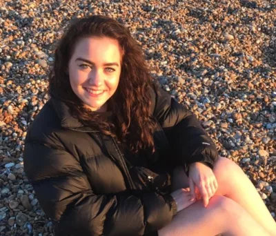 A young woman sits on a pebble beach, smiling for the camera.