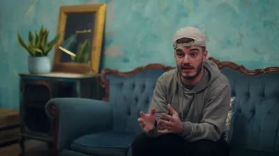 A young man in a backwards cap sits on a blue sofa while gesturing to someone behind the camera.