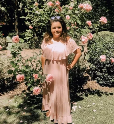 A young woman in a peach dress and sunglasses stands in a rose garden.