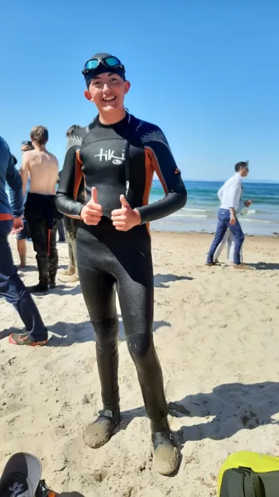 Zac, a teenager, gives two thumbs up while standing in a wetsuit on a sandy beach.