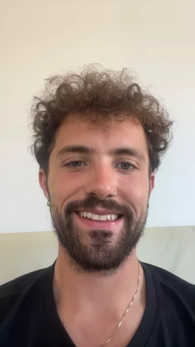 A young man, Miles, with a short beard and curly brown hair, smiles to camera.