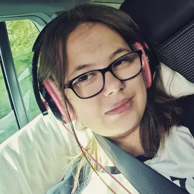 A young woman in glasses and headphones smiles in a selfie in a car.