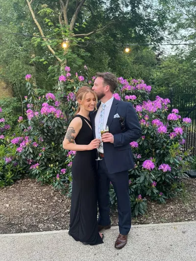 A young couple dressed in formalwear pose before a bush of purple flowers.