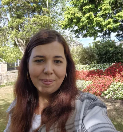 A young woman with long red hair smiles in a selfie from a flowery garden.