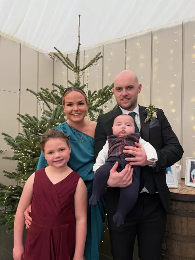 A woman, a man, and their two young child smile in formal wear in front of a Christmas tree.