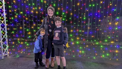A mother and her two sons smile at Christmas time in front of a wall of fairy lights.