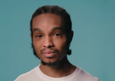 A young black boy stands against a blue backdrop wearing a white t-shirt
