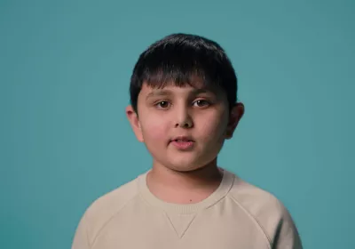 A young Asian boy stands against a blue backdrop wearing a white t-shirt