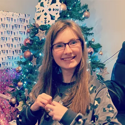 A young woman in glasses smiles before a Christmas tree.