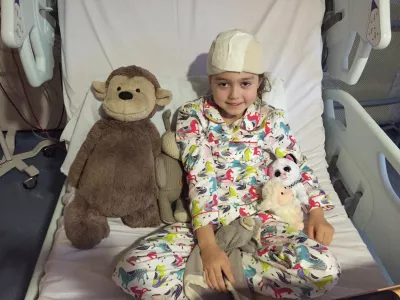 A young girl with a surgery bandage on her head sits with her stuffed teddies.