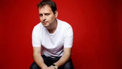 Adam Kay kneels before a red background.