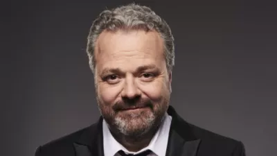 Comedian Hal Cruttenden smiles in a suit against a grey background.