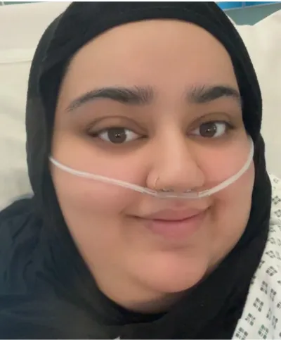 A young woman in a headscarf sits in a hospital bed with a breathing tube.