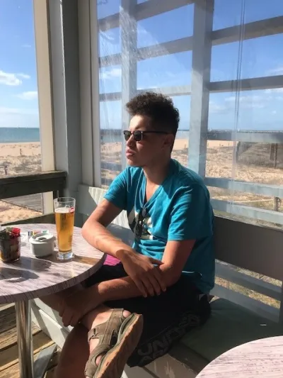 A young man in a blue t-shirt sits with a beer overlooking a beautiful view.