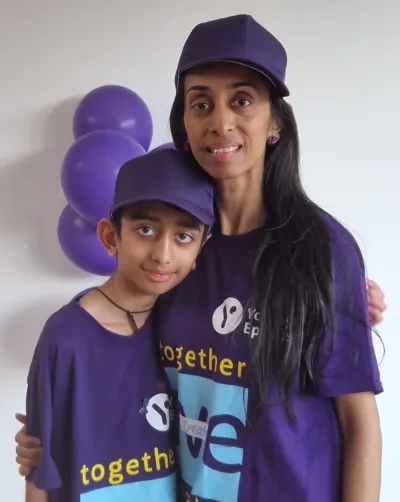 A mother and son in Young Epilepsy clothes smile before purple balloon, wearing Young Epilepsy merchandise.