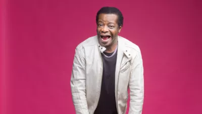 Comedian, Stephen K Amos poses with his mouth open with a silly face on a pink background.