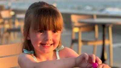 A young girl grins while sitting outdoors in the sunshine.